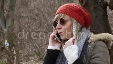 年轻漂亮的女人，戴着红帽子坐在树旁，用手机<strong>说话</strong>